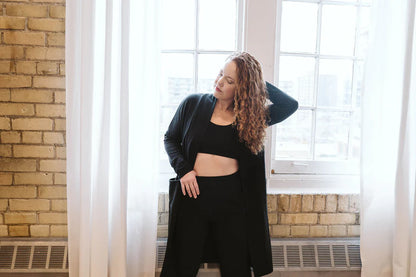 A woman wearing the Courtney Cardigan by Advika in Black, a long pen cardigan with patch pockets, made from tencel/organic eco-fabric, over legggings and a bra. She is standing in front of a window in a brick wall. 